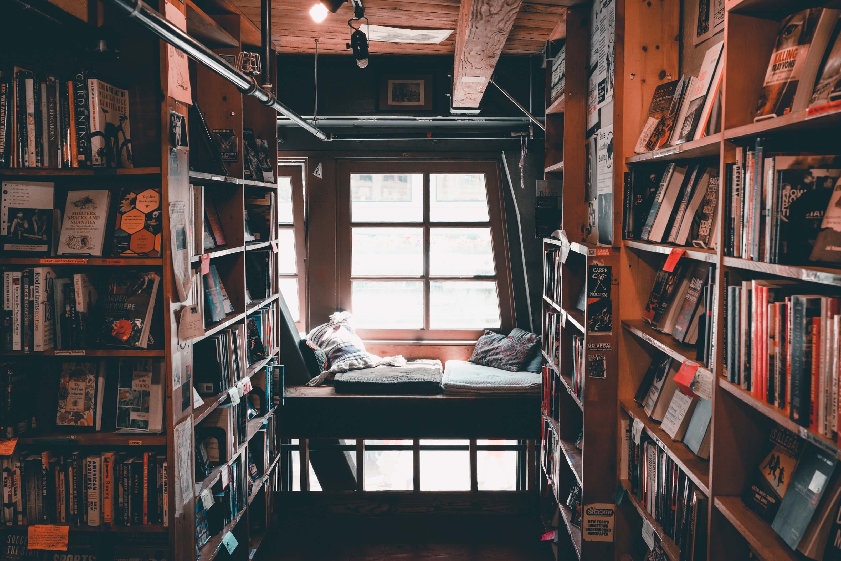 Stock photo by Clay Banks depicting a window seat in between multiple bookshelves filled with stacks of books.